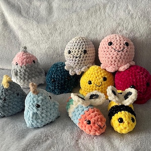 Può includere: Una collezione di peluche all'uncinetto fatti a mano in vari colori e forme. Include polpi, balene e api sorridenti. I peluche sono realizzati con filato morbido e strutturato e presentano occhi di sicurezza neri. La tavolozza dei colori comprende rosa, giallo, blu e rosso.