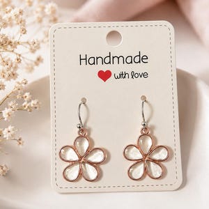 Puede incluir: Un par de pendientes colgantes con forma de flor de cinco pétalos, con bordes de metal en tono oro rosa y centros blancos translúcidos. Los pendientes están sujetos a una tarjeta de presentación blanca con el texto Handmade with love.