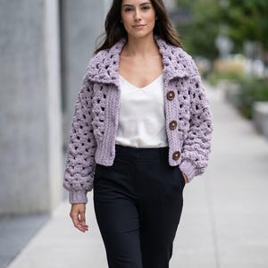 Könnte beinhalten: Eine hellviolette Häkeljacke mit großen Knöpfen. Die Jacke hat einen Kragen und lange Ärmel. Die Person trägt ein weißes Oberteil und eine schwarze Hose. Der Hintergrund ist eine verschwommene Szene im Freien.