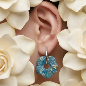 Puede incluir: Un pendiente de aro plateado con un colgante circular en forma de flor en tonos azules y blancos con motas doradas. El colgante tiene una superficie estriada y texturizada, y está suspendido de un aro de plata liso y pulido.