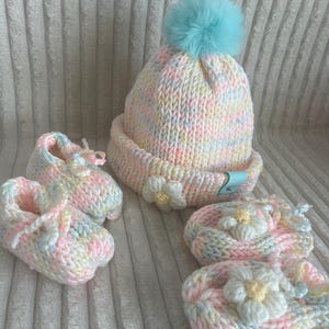 Könnte beinhalten: Ein Set an gestrickten Baby-Accessoires in Pastellfarben. Das Set beinhaltet eine Mütze mit einem blauen Bommel, ein Paar Stiefelchen mit Blumenschmuck und ein zweites Paar Stiefelchen mit Blumenschmuck.