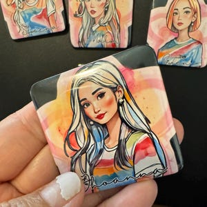 May include: A square button with a watercolor-style illustration of a woman with long blonde hair. She is wearing a rainbow striped shirt and has a pink blush on her cheeks. The text "Joanna" is written in a cursive font at the bottom of the button.