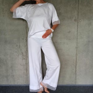 Könnte beinhalten: Weißes zweiteiliges Outfit mit einem locker sitzenden Oberteil und einer weiten Hose. Beide Teile haben eine zarte Spitzenborte an den Ärmeln und Säumen.