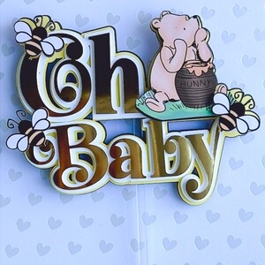 May include: A layered cake topper with the words "Oh Baby" in gold and brown tones, featuring a cartoon bear and bees. The bear is sitting next to a honey pot. The topper is on a clear plastic stick.