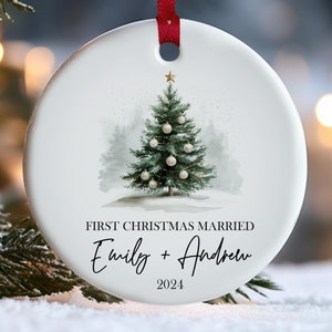 May include: White ceramic Christmas ornament with a watercolor illustration of a Christmas tree and the text "First Christmas Married" with names and the year 2024. A red ribbon is attached for hanging.