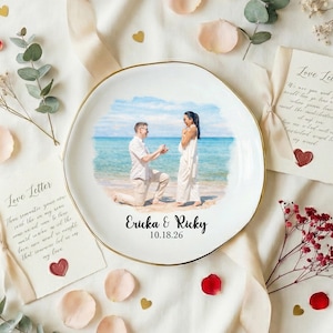 Puede incluir: Un plato de cer&aacute;mica blanco con borde dorado presenta una escena de propuesta en la playa. El plato est&aacute; rodeado de elementos decorativos, como p&eacute;talos de rosa, eucalipto y cartas de amor. Los nombres "Ericka & Ricky" y la fecha "10.18.26" est&aacute;n impresos.