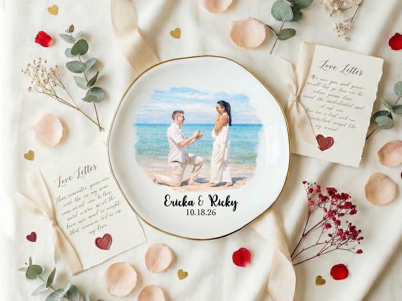 Puede incluir: Un plato de cer&aacute;mica blanco con borde dorado presenta una escena de propuesta en la playa. El plato est&aacute; rodeado de elementos decorativos, como p&eacute;talos de rosa, eucalipto y cartas de amor. Los nombres "Ericka & Ricky" y la fecha "10.18.26" est&aacute;n impresos.