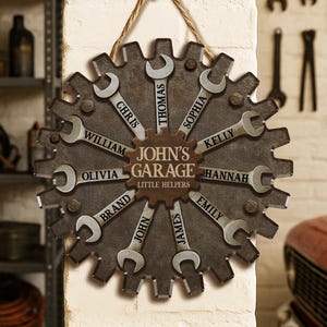 May include: A personalized garage sign shaped like a gear, featuring wrenches with names like Chris, Thomas, and Olivia. The sign reads "John's Garage Little Helpers" and is suspended by a rope.