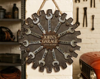 Op de afbeelding: Een gepersonaliseerd garagebord in de vorm van een tandwiel, met sleutels met namen als Chris, Thomas en Olivia. Het bord leest "John's Garage Little Helpers" en is opgehangen met een touw.