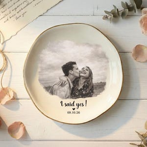 Puede incluir: Un plato de cer&aacute;mica color crema con borde dorado presenta una foto en blanco y negro de una pareja bes&aacute;ndose. El texto "I said yes!" y la fecha 09.10.26 est&aacute;n impresos debajo. El plato est&aacute; rodeado de flores y p&eacute;talos.