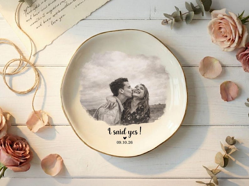 Puede incluir: Un plato de cer&aacute;mica color crema con borde dorado presenta una foto en blanco y negro de una pareja bes&aacute;ndose. El texto "I said yes!" y la fecha 09.10.26 est&aacute;n impresos debajo. El plato est&aacute; rodeado de flores y p&eacute;talos.