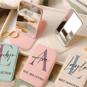 May include: A collection of personalized compact mirrors and small tins. The mirrors are pink and white, with a hinged design. The tins are pastel colors, each with a name and message: "My Reflection", "You Are Loved", and "Best Mom Ever".