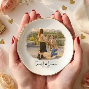 Puede incluir: Un plato de cer&aacute;mica blanco con borde dorado, con una ilustraci&oacute;n en acuarela de una propuesta de matrimonio. El plato est&aacute; personalizado con los nombres "David & Lauren" y la fecha 02.16.26. El plato se sostiene en manos, rodeado de rosas y un joyero.