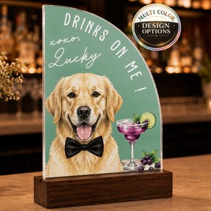 May include: A semi-circular acrylic sign with a dark wood base featuring a golden retriever wearing a black bow tie, a purple cocktail, and the text "DRINKS ON ME!" and "xoxo, Lucky" on a sage green background with a circular "MULTI COLOR DESIGN OPTIONS" badge.