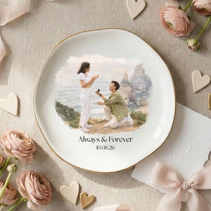 Puede incluir: Un plato de cer&aacute;mica blanca con borde dorado presenta una escena pintada de una propuesta de matrimonio. El plato est&aacute; inscrito con "Always & Forever" y una fecha. La escena est&aacute; rodeada de flores, corazones y una cinta.