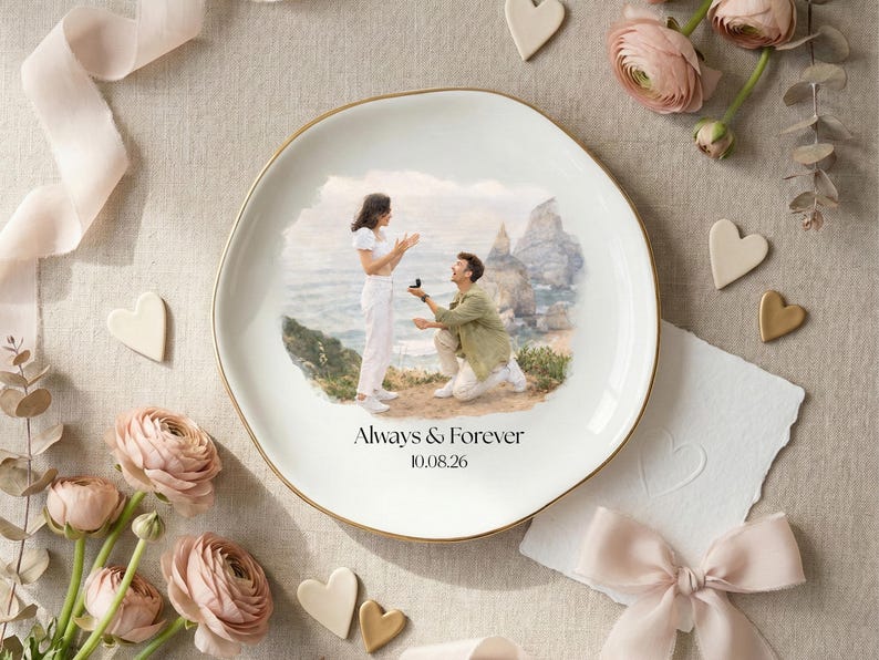 Puede incluir: Un plato de cer&aacute;mica blanca con borde dorado presenta una escena pintada de una propuesta de matrimonio. El plato est&aacute; inscrito con "Always & Forever" y una fecha. La escena est&aacute; rodeada de flores, corazones y una cinta.