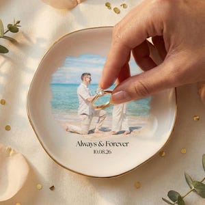 Puede incluir: Un plato de cer&aacute;mica blanco con borde dorado, con una foto de una propuesta de matrimonio en una playa. El plato tiene las palabras "Always & Forever" y la fecha "10.08.26" impresas. Una mano sostiene un anillo de oro.