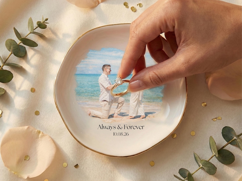 Puede incluir: Un plato de cer&aacute;mica blanco con borde dorado, con una foto de una propuesta de matrimonio en una playa. El plato tiene las palabras "Always & Forever" y la fecha "10.08.26" impresas. Una mano sostiene un anillo de oro.