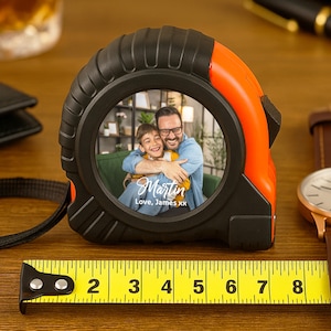 May include: Black and orange tape measure with a photo of a father and child. The tape measure shows measurements up to 8 inches. A brown leather watch and black wallet are also visible.