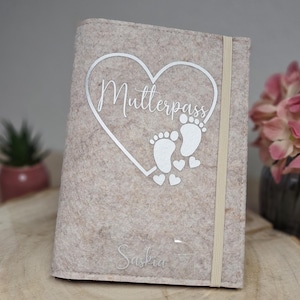 Könnte beinhalten: Ein beigefarbener Filz-Reisepass-Etui mit einem silbernen Herz-Design. Das Herz trägt den Text "Mutterpass" und ein Paar Fußabdrücke im Inneren. Der Name "Saskia" ist unter dem Herzen gedruckt.