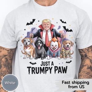 Könnte beinhalten: Hellgraues T-Shirt mit einer Cartoon-Illustration eines als Vampir verkleideten Mannes, der Leinen hält, die an vier Hunden in Halloween-Kostümen befestigt sind. Der Text "JUST A TRUMPY PAW" ist darunter gedruckt.