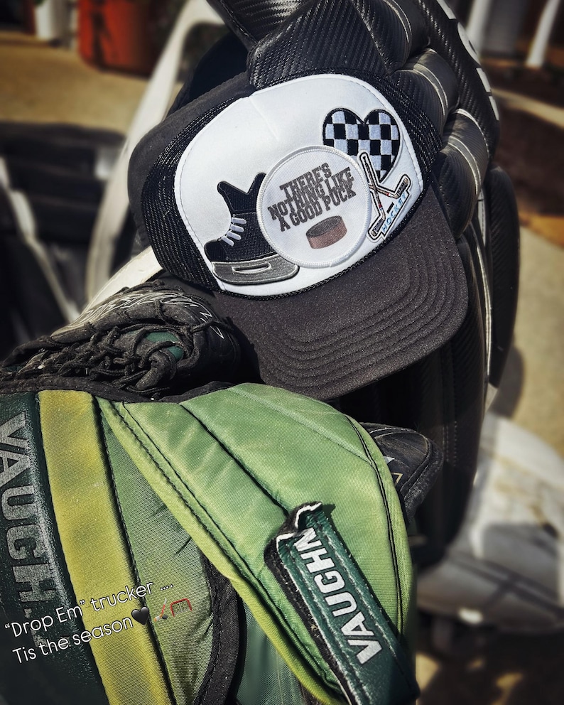 May include: A black and white trucker hat with a hockey-themed patch that says "There's nothing like a good puck." The hat is sitting on top of a green hockey bag with the word "Vaughn" written on it.