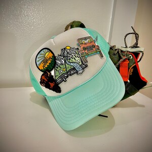 May include: A turquoise and white trucker hat with a patch featuring a mountain scene, a jeep, and the text "Seek Adventure".