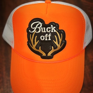 Może przedstawiać: Pomarańczowa i biała siatkowa czapka trucker z czarną naszywką z napisem "Buck off" białymi literami i złotymi porożem.