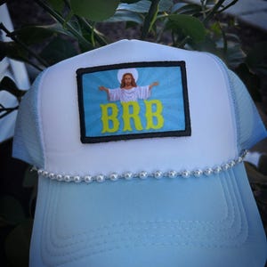 Puede incluir: Una gorra de camionero azul claro con un panel frontal blanco y un ribete de perlas blancas. El panel frontal presenta un parche con una imagen de Jesús con el texto "BRB" en letras amarillas.