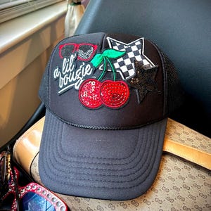 Puede incluir: Gorra de camionero negra con un parche de cereza rojo y verde, un parche de estrella a cuadros blanco y negro y un parche de estrella negra. La gorra también tiene un parche de gafas de sol rojo y negro y el texto "a lil boujie" en blanco.