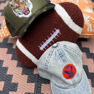 May include: An assortment of caps and a football-shaped pillow. One cap is olive green with a tiger patch, another is denim with a red and blue patch, and the pillow is brown with white stripes.
