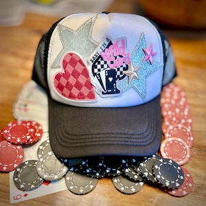 May include: White trucker hat with black mesh back. The hat has several patches including a pink and white checkered heart, a black and white playing card patch with the text "Baby, I'm a Wild Card", and two silver glitter stars.