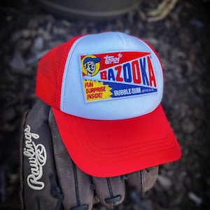 Puede incluir: Una gorra de béisbol roja y blanca con un logotipo de Bazooka Bubble Gum. El logotipo presenta un personaje de dibujos animados y el texto "Fun Surprise Inside!" y "Bazooka Bubble Gum".