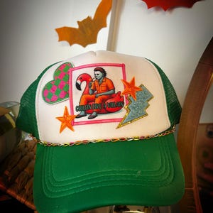 May include: Green and white trucker hat with a variety of colorful patches. The patches include a pink flamingo, a star, a heart, and a patch with a character in an orange jumpsuit sitting on a flamingo float with the text "Chillin' Like a Villain".