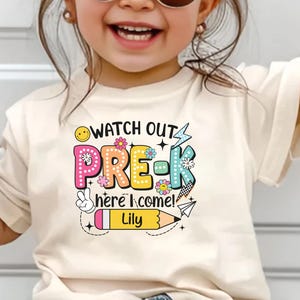 May include: A cream-colored t-shirt with the text "WATCH OUT PRE-K here I come!" in colorful, playful letters. The design includes a smiley face, flowers, a peace sign, and a pencil with the name "Lily".