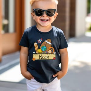 May include: A navy blue t-shirt with a colorful school-themed graphic featuring a rocket, football, globe, and ruler. The name "Noah" is printed on the ruler. The child is wearing sunglasses and white shorts.
