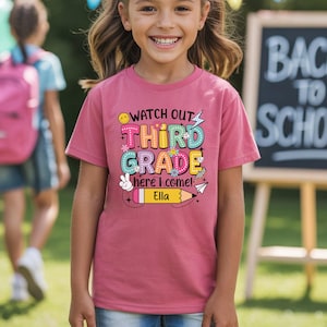 May include: A mauve-colored t-shirt with the words "Watch Out Third Grade Here I Come!" in a colorful, playful font. The name "Ella" is printed below a yellow pencil graphic. The shirt is worn by a smiling child.