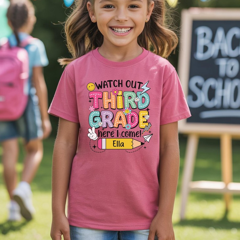 May include: A mauve-colored t-shirt with the words "Watch Out Third Grade Here I Come!" in a colorful, playful font. The name "Ella" is printed below a yellow pencil graphic. The shirt is worn by a smiling child.