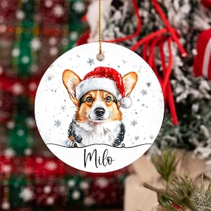 May include: Round Christmas ornament featuring a corgi wearing a Santa hat. The ornament has a white background with snowflakes and the name "Milo" in script. A gold string is attached for hanging.