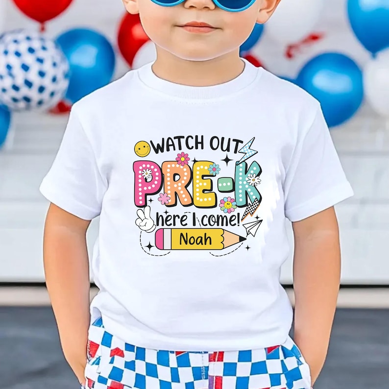 May include: White t-shirt with the text "WATCH OUT PRE-K here I come! Noah" in colorful letters and graphics. The design includes a smiley face, flowers, a peace sign, a pencil, and a paper airplane. The shirt is worn by a child.
