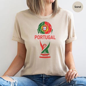 May include: A sand-colored t-shirt featuring a graphic of a soccer ball colored with the green and red of the Portuguese flag, the word PORTUGAL in red text, and a stylized trophy icon with the word PORTUGAL printed at the base of the design.
