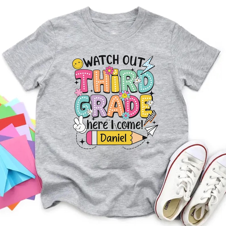 May include: A gray t-shirt with the text "WATCH OUT THIRD GRADE here I come! Daniel" in colorful bubble letters and graphics. The design includes a smiley face, flowers, a peace sign, a pencil, and a paper airplane. White sneakers are visible.