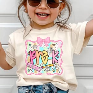 May include: A cream-colored t-shirt featuring a colorful graphic design for pre-kindergarten. The design includes the text "pre-k" in pastel colors, daisies, stars, a pink bow, a ruler, and a pencil.