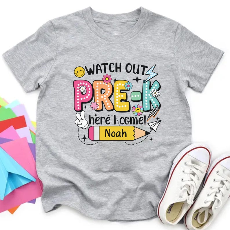 May include: A gray t-shirt with the words "WATCH OUT PRE-K here I come!" in colorful letters and graphics. A pencil with the name "Noah" is also featured. White sneakers are at the bottom right.