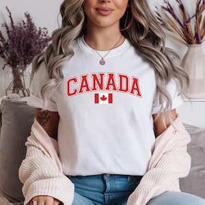 May include: A white short-sleeved t-shirt featuring the word CANADA printed in a bold red collegiate font across the chest, positioned directly above a small, centered graphic of the red and white Canadian national flag with a maple leaf.