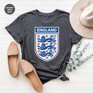 May include: A heather dark gray t-shirt featuring a white shield graphic with the text ENGLAND at the top and three blue lions stacked vertically, surrounded by small red floral accents, with short sleeves and a crew neckline.