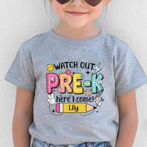 May include: A gray short-sleeved t-shirt with the words "WATCH OUT PRE-K here I come!" in colorful letters and a pencil graphic. The name "Lily" is printed on the pencil.