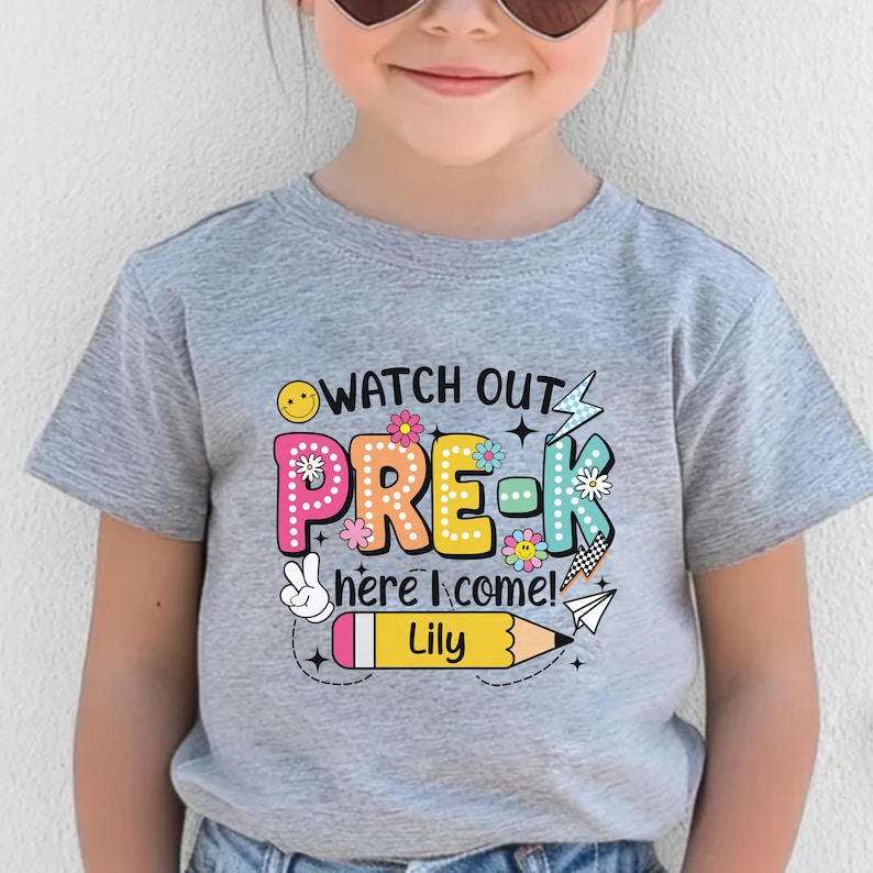 May include: A gray short-sleeved t-shirt with the words "WATCH OUT PRE-K here I come!" in colorful letters and a pencil graphic. The name "Lily" is printed on the pencil.