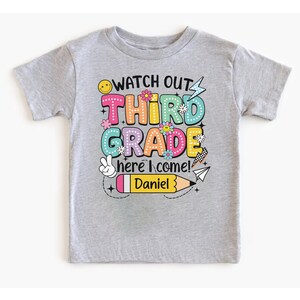 May include: A light gray t-shirt with the words "WATCH OUT THIRD GRADE here I come!" in colorful bubble letters. A yellow pencil with the name "Daniel" is also featured on the shirt. The shirt is designed for a child.
