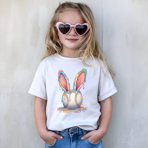 May include: A light-colored t-shirt featuring a baseball with colorful, dripping bunny ears. The baseball has red stitching. The shirt is worn by a person wearing pink heart-shaped sunglasses.